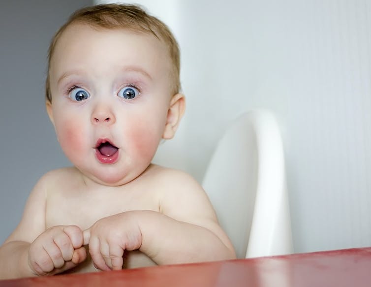 A baby makes a comical 'shocked' face, with wide eyes and an open mouth.