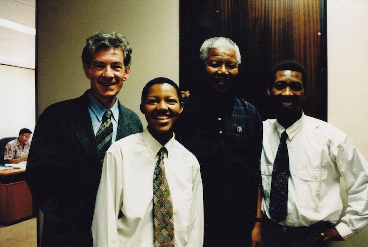 Four people of differing ages pose with an elderly man in a nondescript office setting. They smile at the camera.